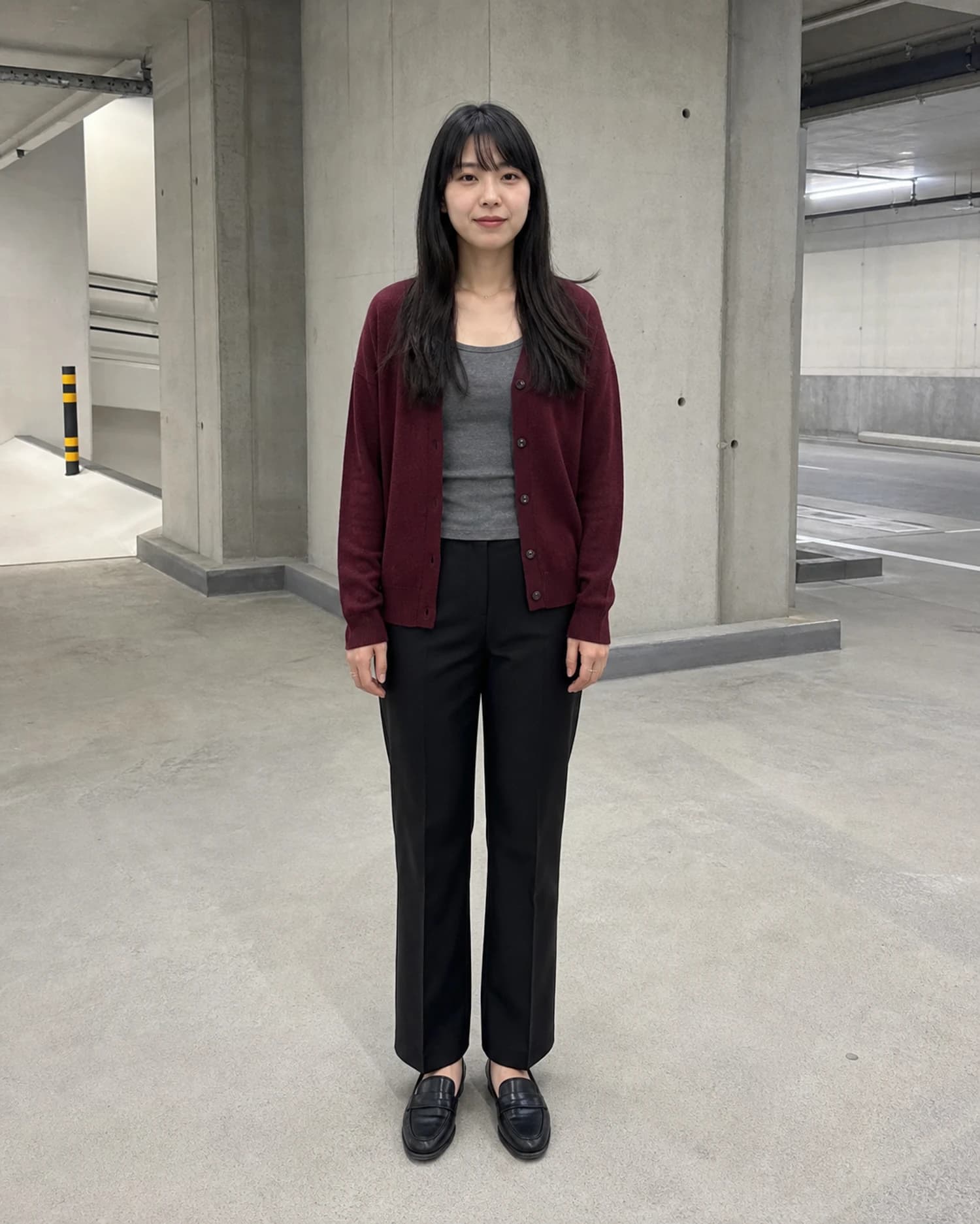 一名成年人物穿酒红色开衫、灰色上衣、黑色长裤和乐福鞋站在混凝土车库中的全身人像