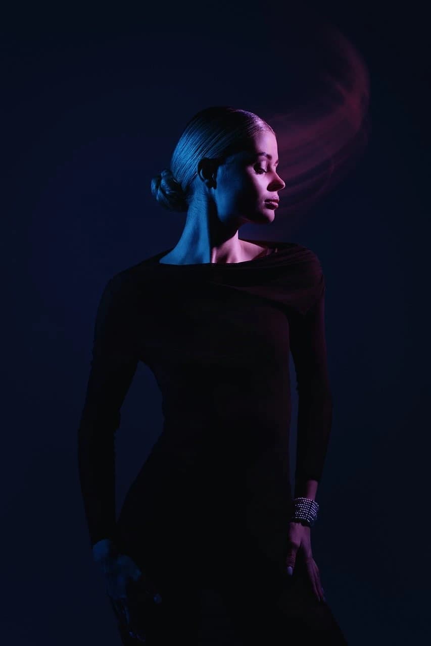 Retrato de estudio de perfil de un adulto con atuendo oscuro, iluminación cian y magenta sobre fondo casi negro