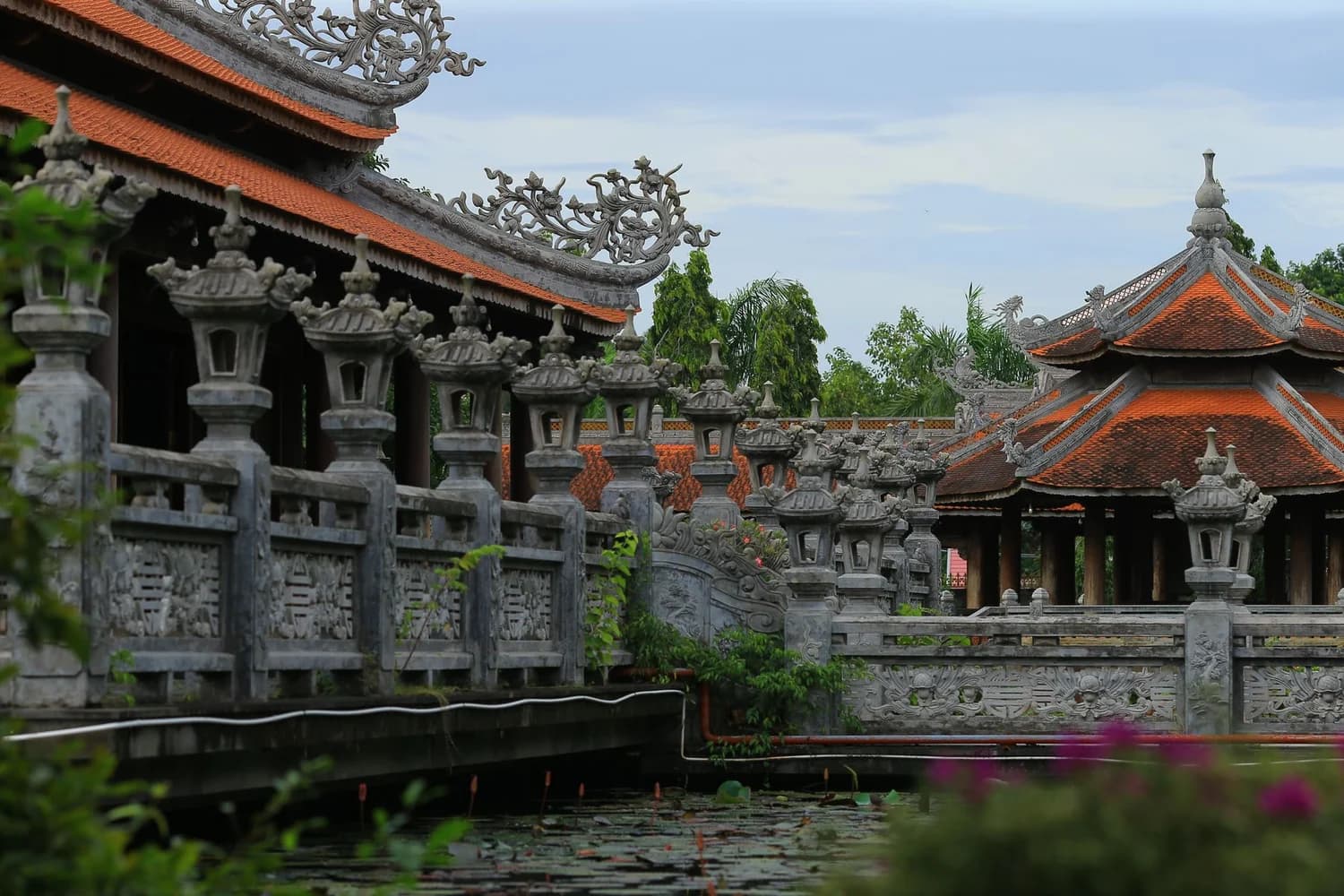 Fotografía real de arquitectura tradicional asiática con tejados de teja, postes de linterna, barandillas y un estanque que refleja el paisaje