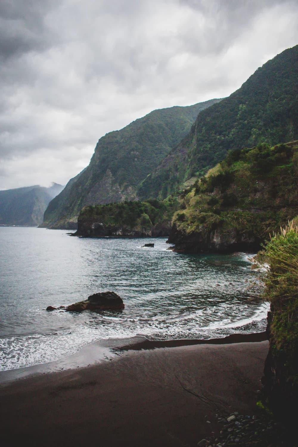 Fotografía original de paisaje costero vertical con montaña oscura a la derecha, agua a la izquierda, playa curva y cielo nublado
