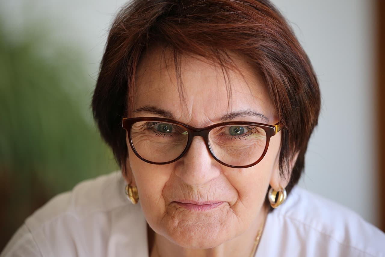 Primer plano de retrato de mujer mayor con cabello corto castaño, gafas redondeadas oscuras, pendientes de aro y blusa blanca mirando directamente a la cámara
