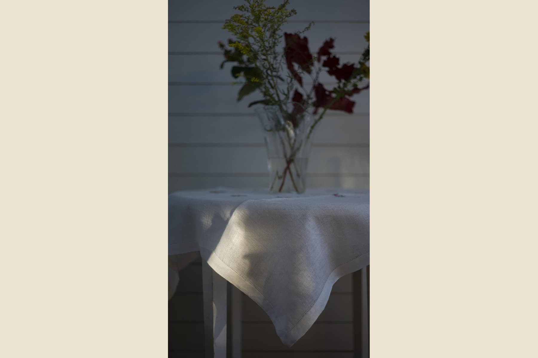 Low-light vertical still-life photo of a clear glass vase with crossed stems, dark red flowers, small yellow blooms, and a white tablecloth against horizontal wall boards