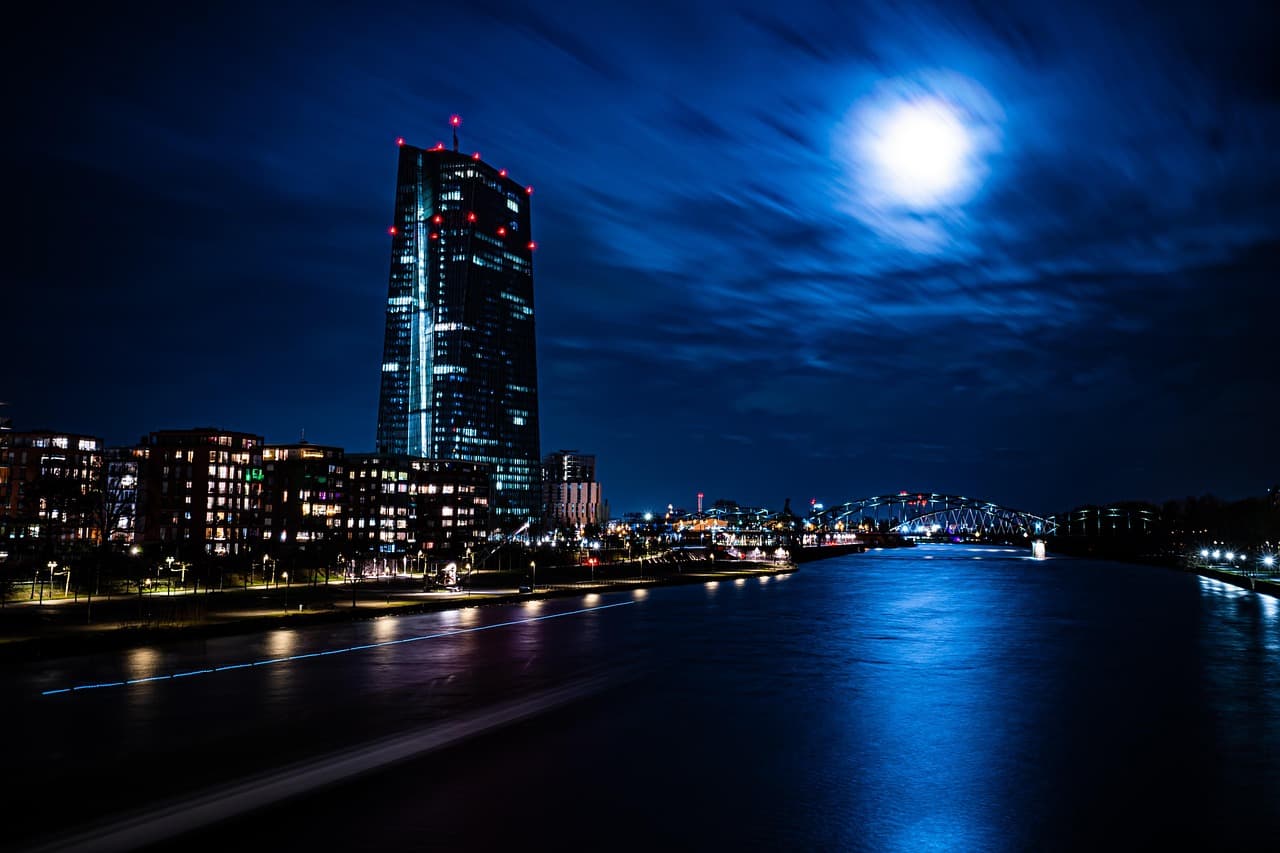 Blue night river scene with an illuminated tower on the left, low arched bridge on the right, and bright moon above calm water
