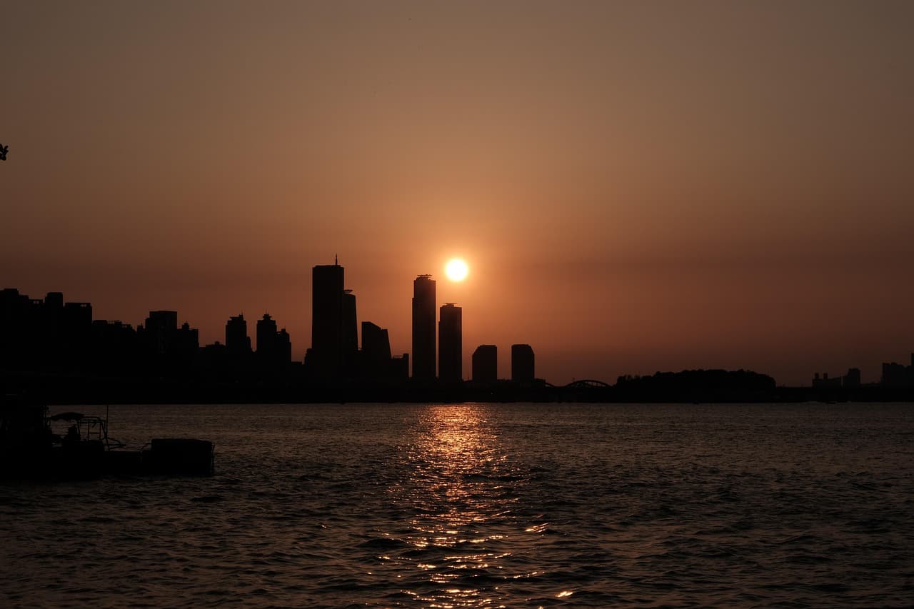 Foto original de un atardecer naranja sobre un horizonte de ciudad oscuro y un río, con el sol encima de las torres centrales y un reflejo brillante que llega hasta la cámara