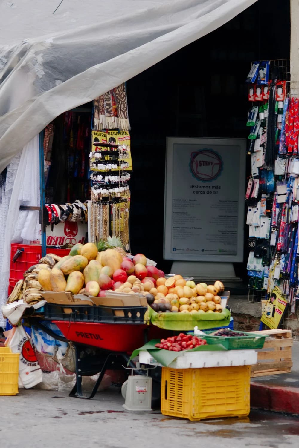 Foto original de un puesto de mercado callejero con toldo inclinado, expositores de frutas, productos colgados y entrada oscura detrás de la mercancía